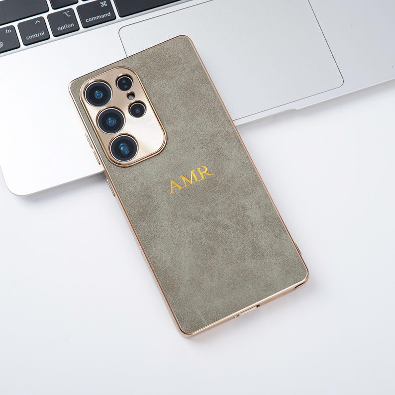 May include: A sage green phone case with gold trim and a gold camera module. The case features the initials "AMR" in gold lettering. The phone is on a white surface next to a laptop.