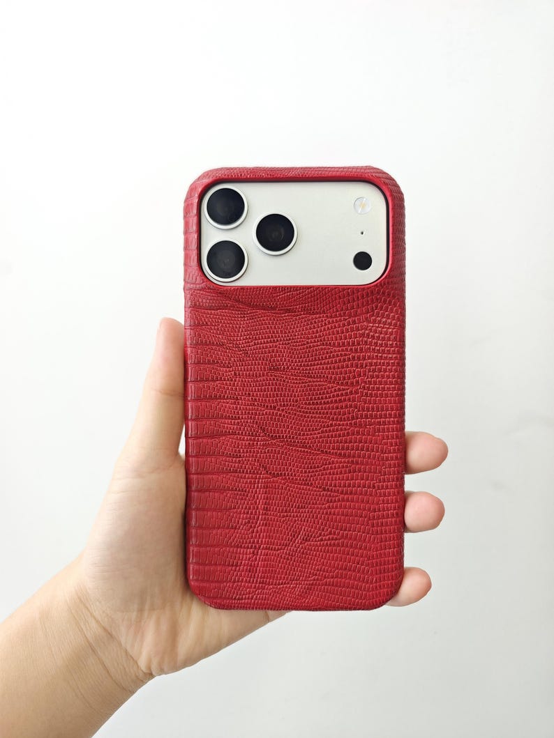 May include: A smartphone case with a textured red finish resembling lizard skin. The case features a rectangular cutout at the top to accommodate the three camera lenses and the flash module of the device, which has a white back panel.