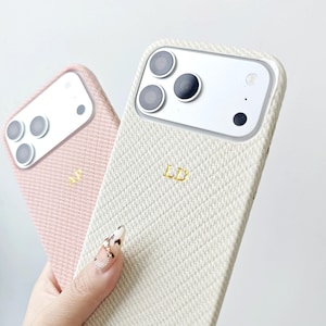 May include: Two smartphone cases with a textured, diagonal weave pattern. One case is light pink with the gold initials AJ, and the other is cream-colored with the gold initials LD. Both cases feature a cutout for a triple-lens camera system.
