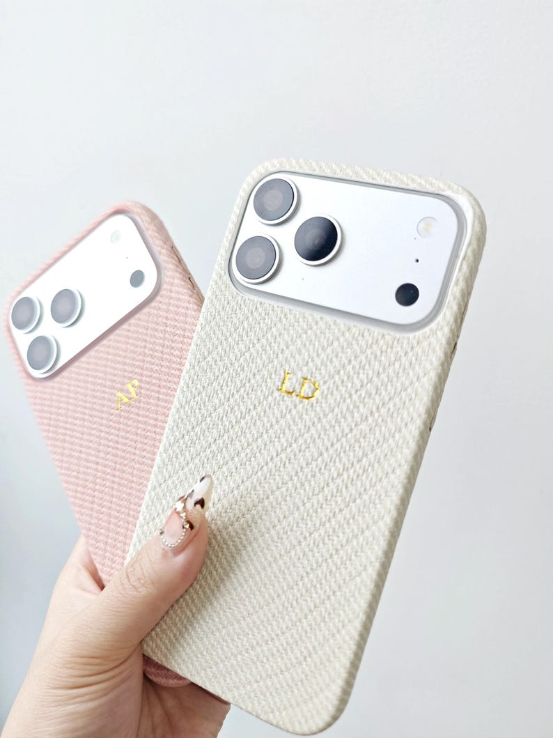 May include: Two smartphone cases with a textured, diagonal weave pattern. One case is light pink with the gold initials AJ, and the other is cream-colored with the gold initials LD. Both cases feature a cutout for a triple-lens camera system.