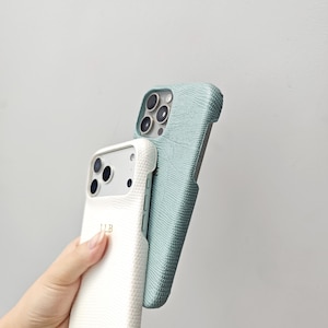 May include: Two smartphone cases with a textured lizard-skin pattern. One case is white with the gold-coloured initials LLB printed on the back, and the other case is a muted sage green. Both cases feature cutouts for the camera lens array.