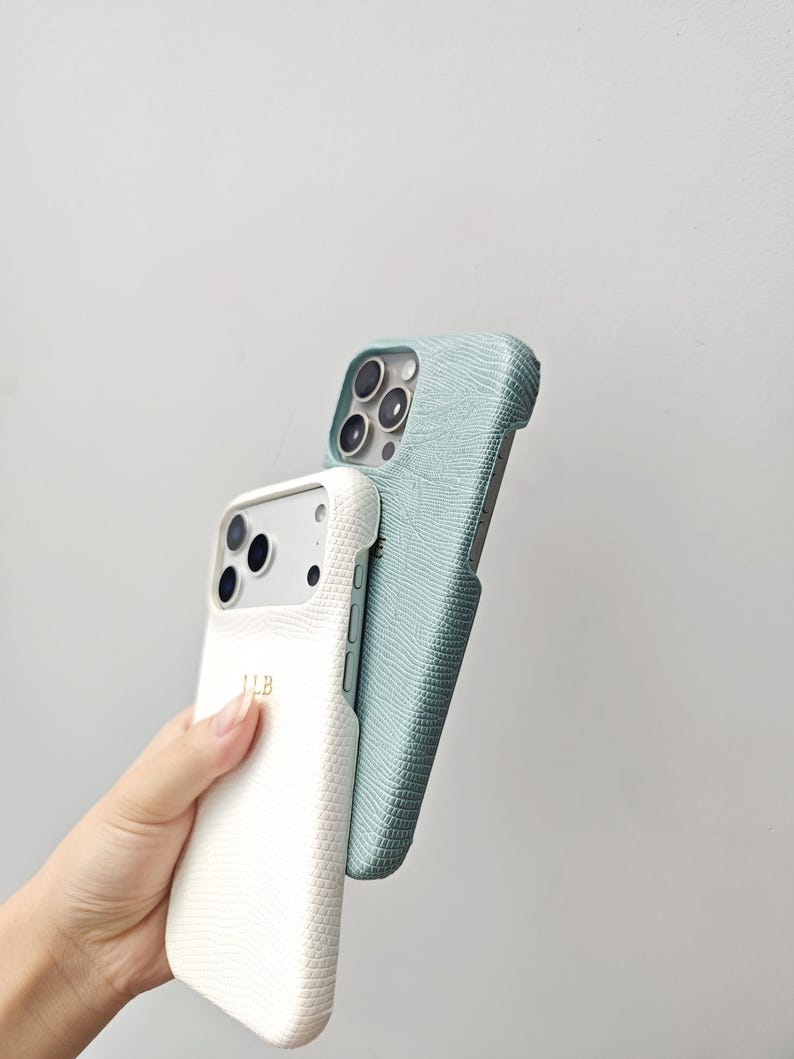 May include: Two smartphone cases with a textured lizard-skin pattern. One case is white with the gold-coloured initials LLB printed on the back, and the other case is a muted sage green. Both cases feature cutouts for the camera lens array.