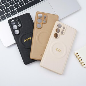 May include: Three personalised phone cases in black, tan, and cream. Each case has a circular design with gold initials. The cases are on a white surface near a laptop.