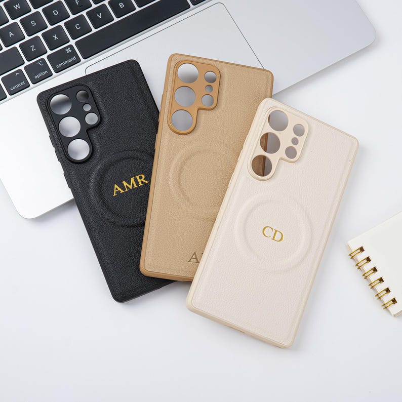 May include: Three personalised phone cases in black, tan, and cream. Each case has a circular design with gold initials. The cases are on a white surface near a laptop.