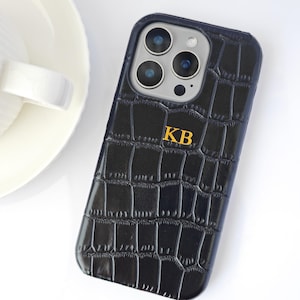 Peut inclure: Coque de t&eacute;l&eacute;phone noire avec une texture peau de crocodile et les initiales dor&eacute;es "KB". L'&eacute;tui a un bord bleu fonc&eacute; et un module cam&eacute;ra avec plusieurs lentilles. Le fond est blanc, avec une partie d'une assiette en c&eacute;ramique blanche visible.