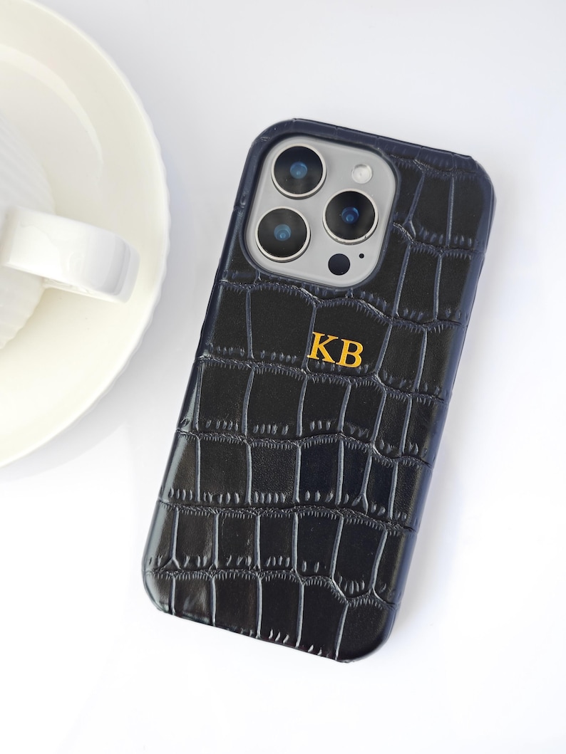 Peut inclure: Coque de t&eacute;l&eacute;phone noire avec une texture peau de crocodile et les initiales dor&eacute;es "KB". L'&eacute;tui a un bord bleu fonc&eacute; et un module cam&eacute;ra avec plusieurs lentilles. Le fond est blanc, avec une partie d'une assiette en c&eacute;ramique blanche visible.