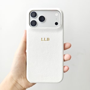 May include: A white smartphone case with a textured, lizard-skin pattern and the gold-coloured letters LLB embossed in the centre. The case features a large rectangular cutout at the top for the camera lenses and flash, showing the phone's silver back.