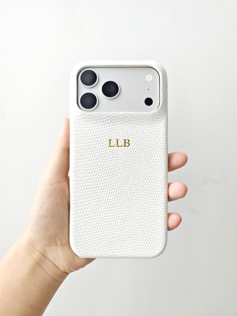 May include: A white smartphone case with a textured, lizard-skin pattern and the gold-coloured letters LLB embossed in the centre. The case features a large rectangular cutout at the top for the camera lenses and flash, showing the phone's silver back.