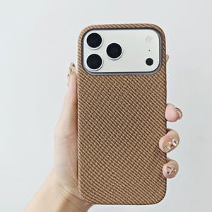 May include: A smartphone case featuring a textured, brown, diagonal herringbone fabric pattern. The case has a rectangular cutout at the top to accommodate a triple-lens camera system and a flash, with a smooth, light-colored border around the lenses.