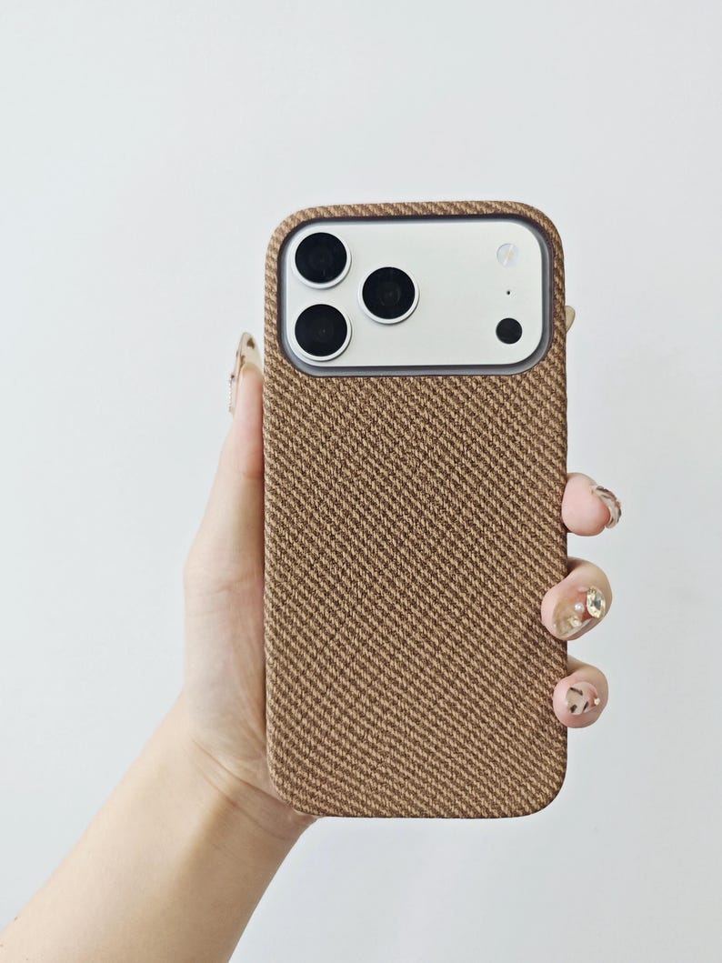 May include: A smartphone case featuring a textured, brown, diagonal herringbone fabric pattern. The case has a rectangular cutout at the top to accommodate a triple-lens camera system and a flash, with a smooth, light-colored border around the lenses.