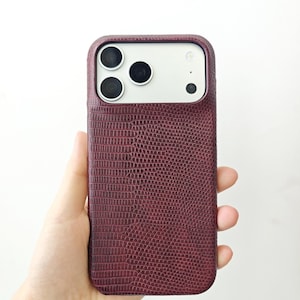 May include: A smartphone case with a deep burgundy, textured lizard-skin pattern. The case features a rectangular cutout at the top to accommodate the three camera lenses and the flash module of the phone, maintaining a sleek and uniform profile.
