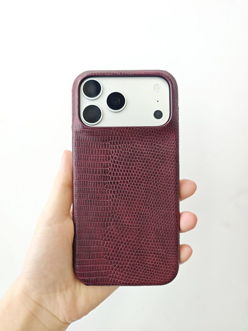 May include: A smartphone case with a deep burgundy, textured lizard-skin pattern. The case features a rectangular cutout at the top to accommodate the three camera lenses and the flash module of the phone, maintaining a sleek and uniform profile.
