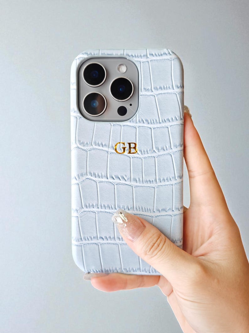 Peut inclure: Coque de t&eacute;l&eacute;phone bleu clair avec une texture imitation peau de crocodile. Le bo&icirc;tier porte un monogramme "GB" dor&eacute;. Le t&eacute;l&eacute;phone a un module cam&eacute;ra avec trois objectifs et un flash. La coque est tenue par une main.