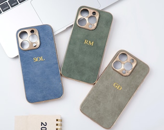 Op de afbeelding: Drie gepersonaliseerde telefoonhoesjes in blauw, groen en grijs, elk met goudkleurige accenten en initialen. De hoesjes zijn gerangschikt op een wit oppervlak in de buurt van een laptop en een klein notitieboekje met het jaar 2025 erop.