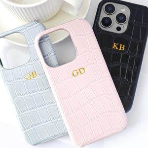 Peut inclure: Trois coques de t&eacute;l&eacute;phone personnalis&eacute;es en bleu clair, rose et noir, chacune avec un monogramme dor&eacute;. Les coques pr&eacute;sentent un motif textur&eacute; en forme de crocodile. Une tasse et une soucoupe blanches sont en arri&egrave;re-plan.