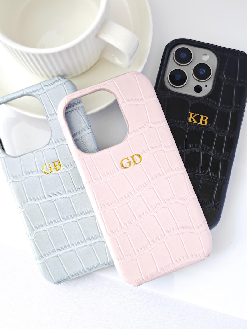 Peut inclure: Trois coques de t&eacute;l&eacute;phone personnalis&eacute;es en bleu clair, rose et noir, chacune avec un monogramme dor&eacute;. Les coques pr&eacute;sentent un motif textur&eacute; en forme de crocodile. Une tasse et une soucoupe blanches sont en arri&egrave;re-plan.