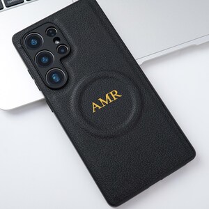 May include: Black leather phone case with the initials "AMR" in gold lettering. The case has a textured surface and a circular indentation. The phone case is on a silver laptop.