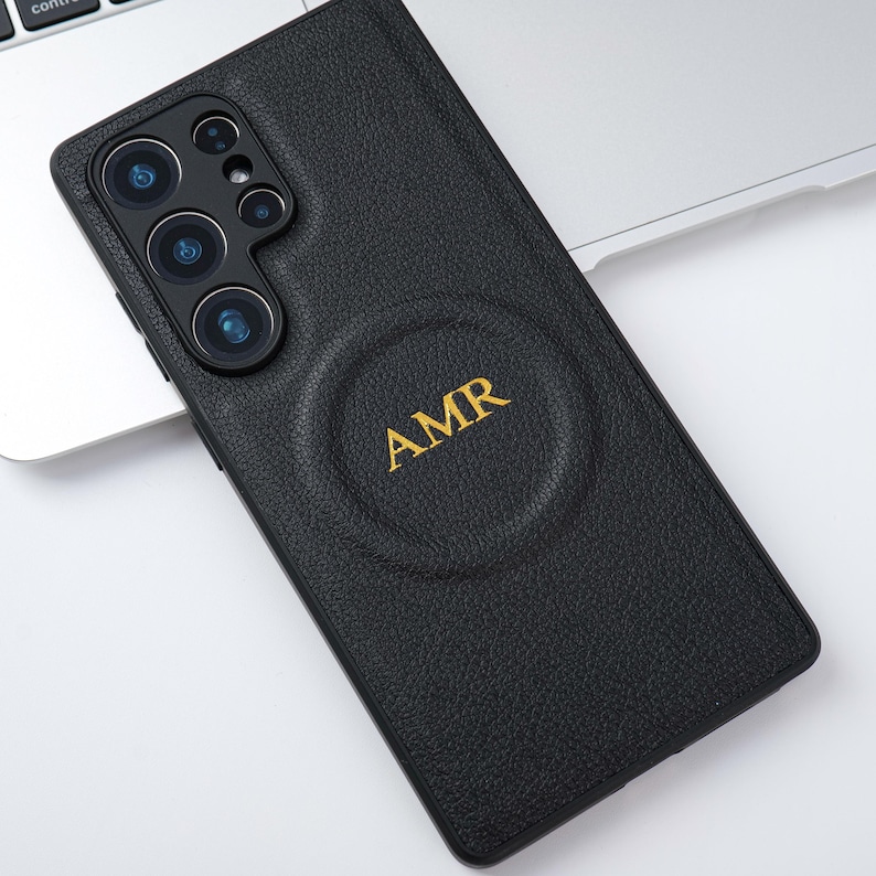 May include: Black leather phone case with the initials "AMR" in gold lettering. The case has a textured surface and a circular indentation. The phone case is on a silver laptop.