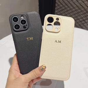 Może przedstawiać: Dwa skórzane etui na telefon, jedno czarne i jedno kremowe, każde ze złotym monogramem. Czarne etui ma inicjały "T.M", a kremowe "A.M". Etui mają teksturowane wykończenie.