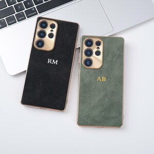 May include: Two smartphone cases with gold camera surrounds and trim. One case is black with the initials "RM" in white, the other is green with the initials "AB" in gold. The cases are on a white surface.