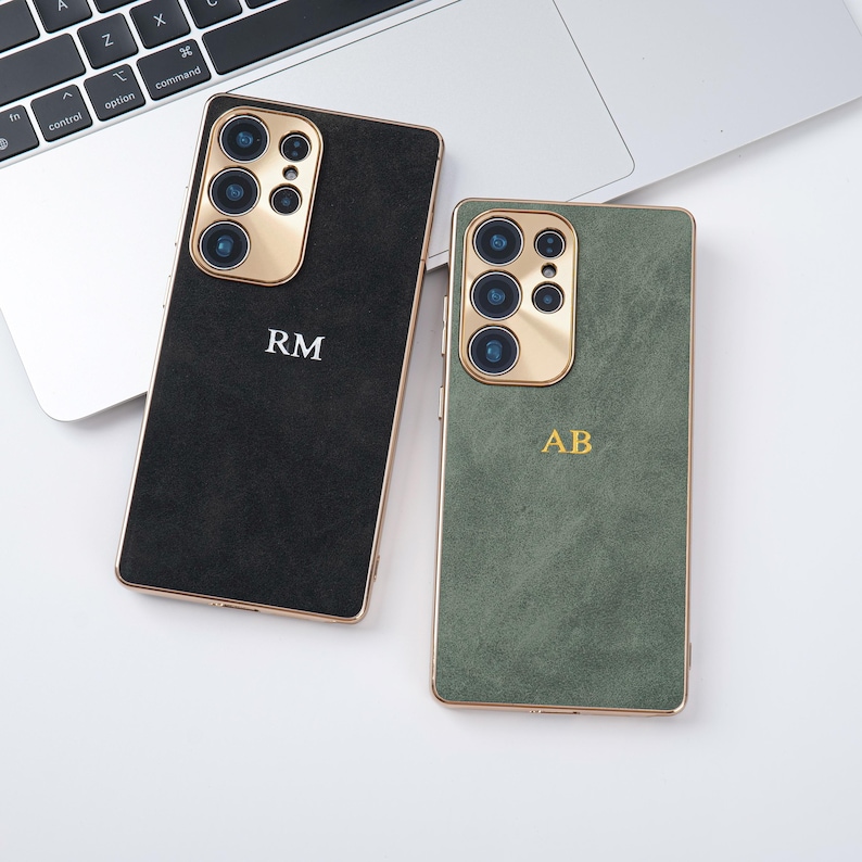 May include: Two smartphone cases with gold camera surrounds and trim. One case is black with the initials "RM" in white, the other is green with the initials "AB" in gold. The cases are on a white surface.
