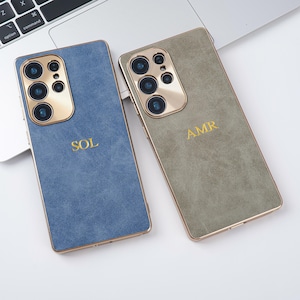 Puede incluir: Dos fundas para smartphone, una azul y otra verde oliva, con contornos y bordes de cámara dorados. La funda azul tiene las letras doradas "SOL", y la funda verde oliva tiene las letras "AMR". Las fundas están sobre una superficie blanca.