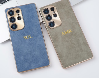May include: Two smartphone cases, one blue and one olive green, with gold-colored camera surrounds and trim. The blue case has the gold-colored letters "SOL", and the olive green case has the letters "AMR". The cases are on a white surface.