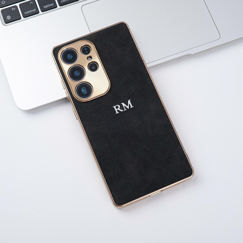 May include: Black smartphone case with gold camera surround and trim. The case features the initials "RM" in white. The phone is on a white surface next to a laptop.