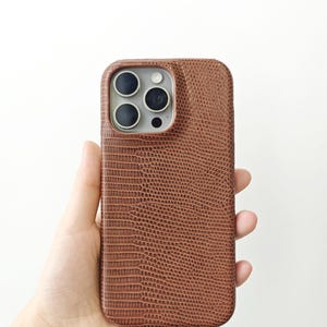May include: A brown smartphone case with a textured lizard skin pattern. The case features a rectangular cutout at the top left corner for the camera lenses, exposing the silver-coloured camera module and three circular black lens openings.