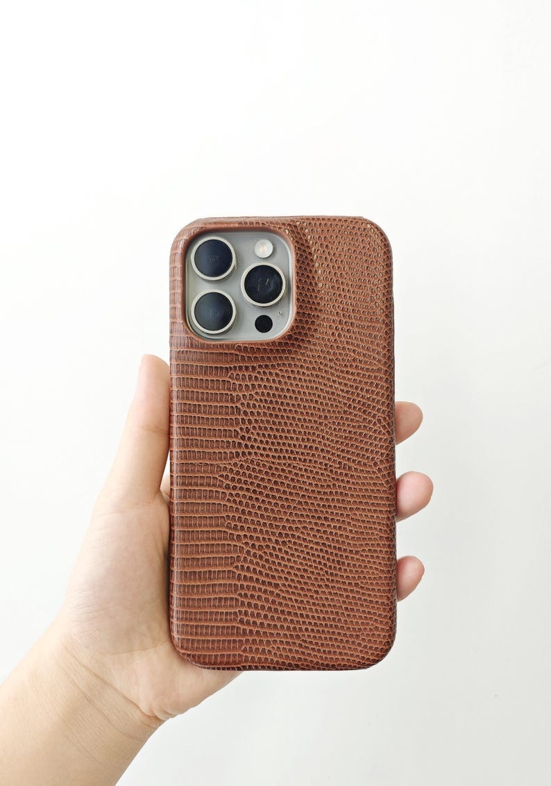 May include: A brown smartphone case with a textured lizard skin pattern. The case features a rectangular cutout at the top left corner for the camera lenses, exposing the silver-coloured camera module and three circular black lens openings.