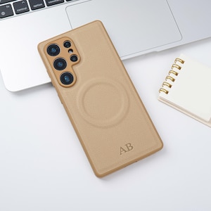 May include: A tan phone case with a textured finish and a circular design on the back. The case features a camera cutout and the initials "AB" are engraved at the bottom. It rests on a white surface beside a laptop and a small notepad.