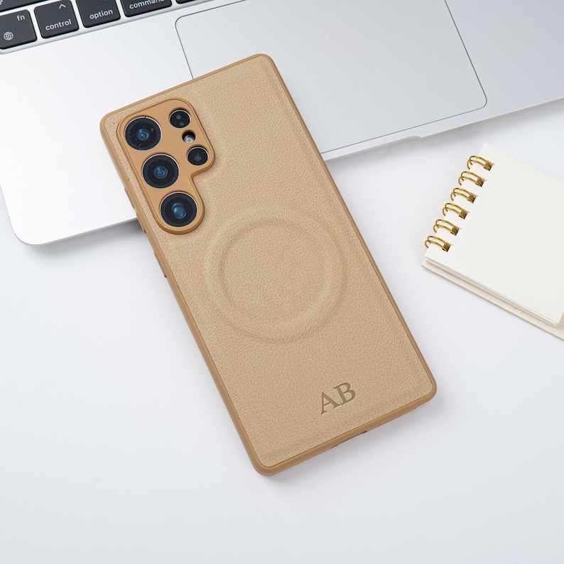 May include: A tan phone case with a textured finish and a circular design on the back. The case features a camera cutout and the initials "AB" are engraved at the bottom. It rests on a white surface beside a laptop and a small notepad.