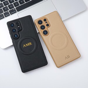 May include: Two phone cases, one black and one tan, are shown. The black case has the initials "AMR" in gold lettering, while the tan case has "AB". Both cases feature a circular design and are placed on a white surface near a laptop.
