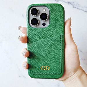 May include: Green leather phone case with a pocket and gold initials. The case has a textured surface and a rectangular shape. The phone case is designed to fit a smartphone.