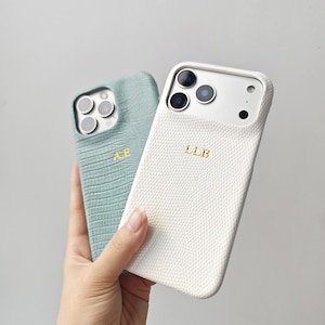 May include: Two phone cases with a textured reptile skin pattern. One case is sage green with the gold embossed initials A.E., and the other is white with the gold embossed initials L.L.B. Both cases feature cutouts for the camera lenses.