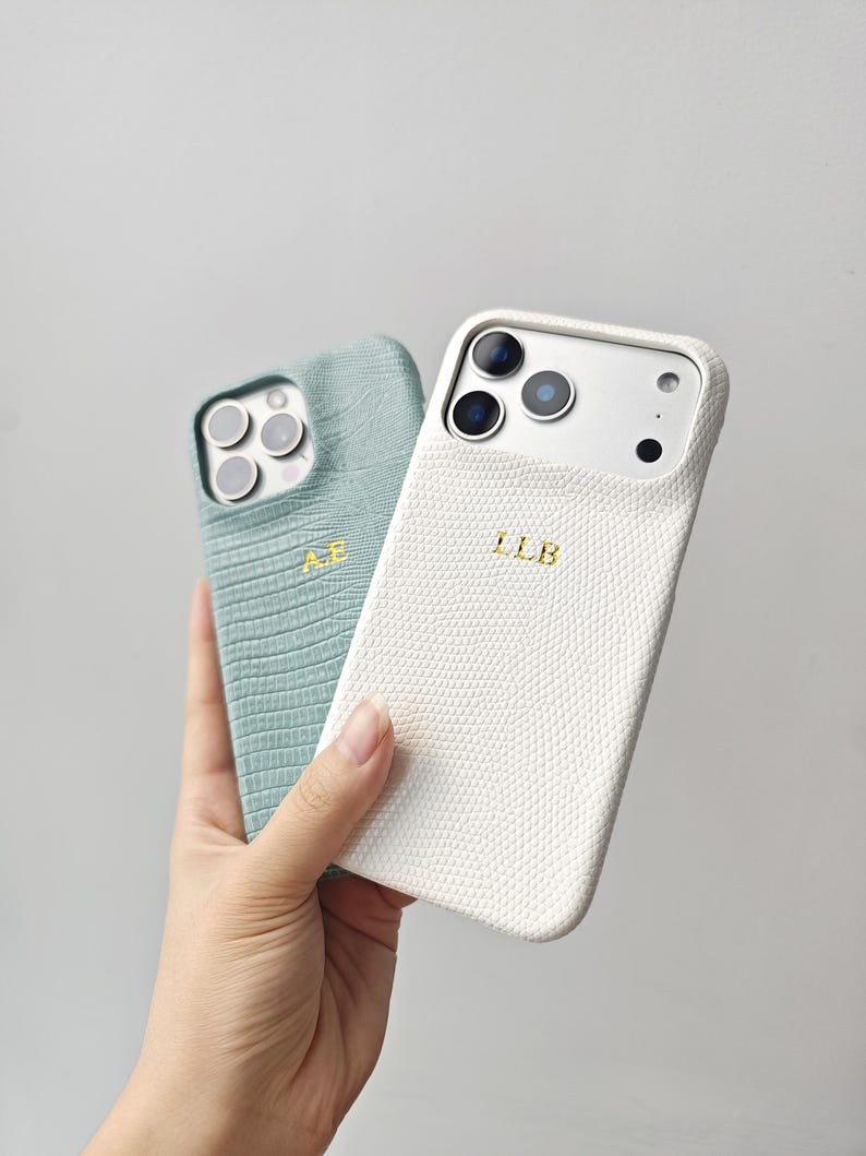 May include: Two phone cases with a textured reptile skin pattern. One case is sage green with the gold embossed initials A.E., and the other is white with the gold embossed initials L.L.B. Both cases feature cutouts for the camera lenses.