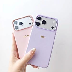 May include: Two smartphone cases in pastel pink and light purple. The pink case features the gold embossed letters LLB, while the purple case displays the gold embossed letters ME. Both cases have a raised camera frame and a smooth finish.