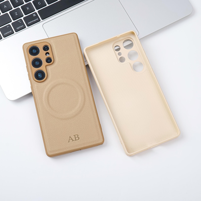 May include: Two phone cases, one tan and one cream, are shown on a white surface near a laptop. The tan case has a circular design and the initials "AB" engraved. The cream case is empty, showing the interior design.