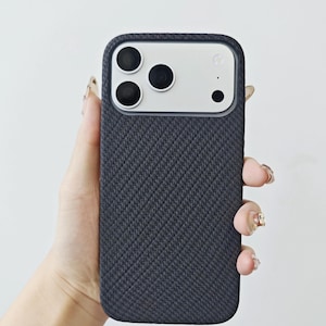 May include: A dark charcoal gray smartphone case with a textured, diagonal woven fabric pattern. The case features a rectangular cutout at the top, revealing a silver camera module with three circular lenses and a small flash component.