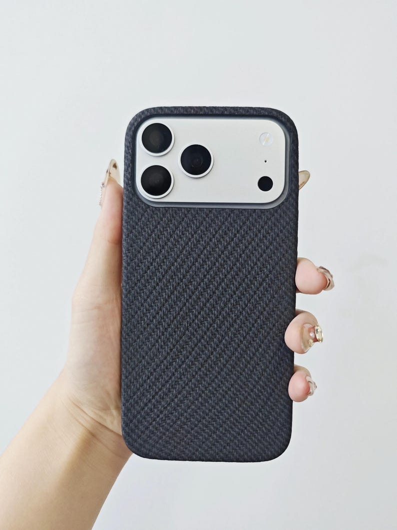 May include: A dark charcoal gray smartphone case with a textured, diagonal woven fabric pattern. The case features a rectangular cutout at the top, revealing a silver camera module with three circular lenses and a small flash component.