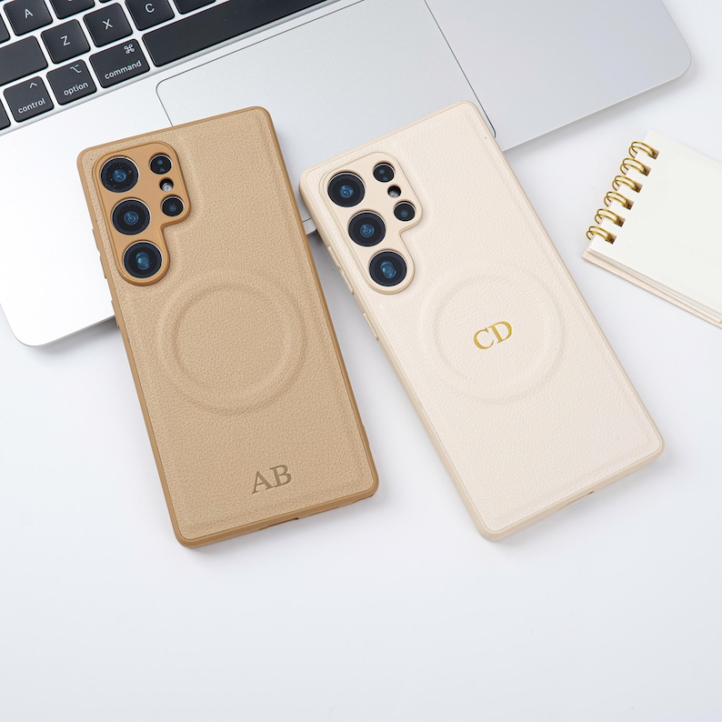 May include: Two phone cases are shown. One is tan with the initials "AB" and the other is off-white with the initials "CD". Both cases have a circular design. A laptop and a small notepad are also visible.