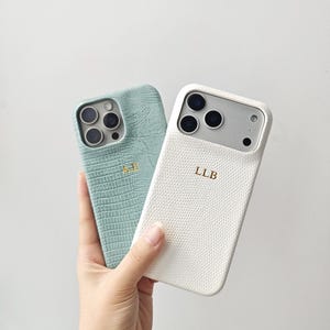 May include: Two smartphone cases with a textured, reptile-like pattern. One case is sage green with the gold embossed initials AE, and the other is white with the gold embossed initials LLB. Both cases feature cutouts for the camera lenses.