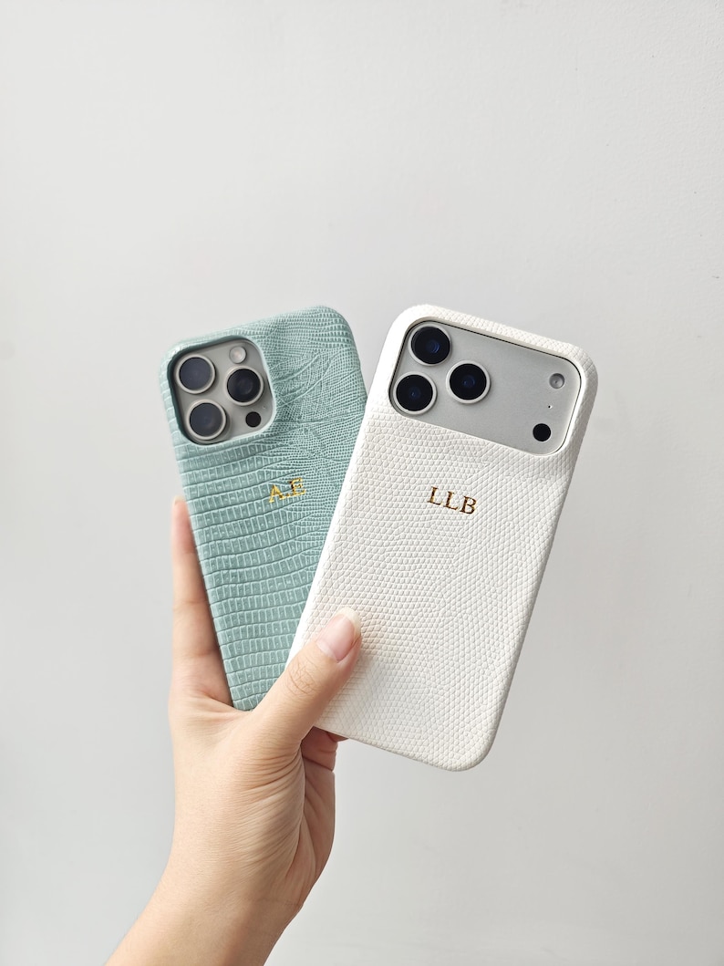 May include: Two smartphone cases with a textured, reptile-like pattern. One case is sage green with the gold embossed initials AE, and the other is white with the gold embossed initials LLB. Both cases feature cutouts for the camera lenses.