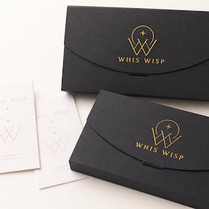 May include: Two rectangular black boxes with a curved flap closure, each featuring a gold foil logo of a circle, a cross, and the text WHIS WISP. Two white rectangular cards are positioned to the left, also displaying the same gold logo and text.