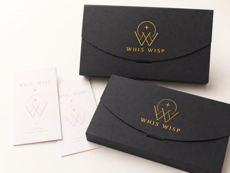 May include: Two rectangular black boxes with a curved flap closure, each featuring a gold foil logo of a circle, a cross, and the text WHIS WISP. Two white rectangular cards are positioned to the left, also displaying the same gold logo and text.