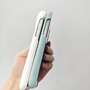 May include: Two smartphone cases with a textured, embossed pattern resembling reptile skin. One case is white and the other is a light blue-green colour. Both cases feature cutouts for side buttons and are shown from a side profile view.
