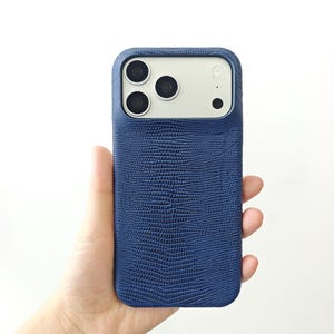 May include: A dark blue smartphone case with a textured, lizard-skin pattern. The case features a rectangular cutout at the top to accommodate a triple-lens camera system and a flash, revealing the light-coloured back of the phone underneath.