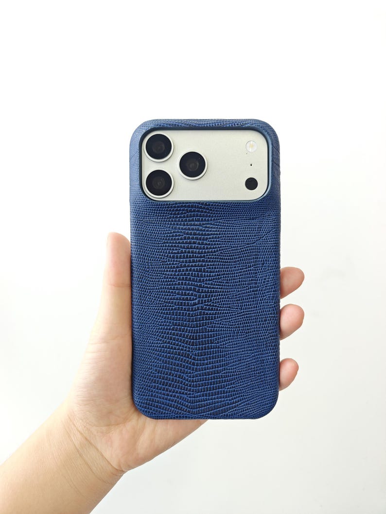 May include: A dark blue smartphone case with a textured, lizard-skin pattern. The case features a rectangular cutout at the top to accommodate a triple-lens camera system and a flash, revealing the light-coloured back of the phone underneath.