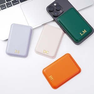 May include: A collection of colorful, rectangular phone wallets in lavender, pink, green, and orange. Each wallet features gold-colored initials. One green wallet is attached to a black phone. The wallets are on a white surface.