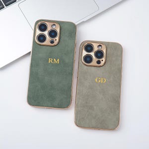 May include: Two olive green phone cases with gold-colored trim and camera surrounds. Each case has a gold monogram, one with "RM" and the other with "GD". The cases are on a white surface near a laptop.